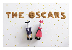 Champagne hold back party favors with tuxedo and dress and mustache and lipstick and popcorn and a voting ballot with pencil on a white fare with confetti and cake truffles in a box with The Oscars written in sugar crystals